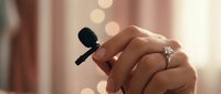 Hands of a woman wearing a diamond engagement ring holding a small lavalier microphone with soft bokeh lights in the background.