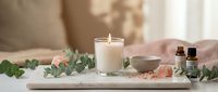 Spa wellness setup with burning candle, essential oils, eucalyptus leaves and pink salt crystals on white tray for relaxation and aromatherapy treatment.