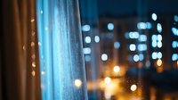 Dreamy evening cityscape through sheer curtain with soft bokeh lights creating magical atmosphere, window view with defocused urban nightscape.