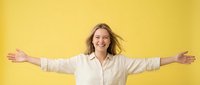 Young woman with shoulder-length blonde hair, Caucasian ethnicity, wearing cream button-up shirt, arms spread wide in celebratory pose against bright yellow background. Suitable for success, happiness, freedom, and positive lifestyle concepts.