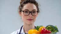 Young female healthcare professional in white coat and glasses holding fresh vegetables and fruits, promoting healthy diet and nutrition counseling concept.