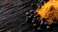 Ground turmeric powder heap and scattered black peppercorns on dark wooden rustic background, macro food photography showing spice texture and details.