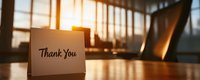 Thank you card on wooden desk in bright modern office with warm sunlight streaming through large windows creating professional gratitude atmosphere.