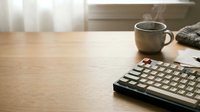 Wooden desk workspace with mechanical keyboard, steaming mug, and natural light from window, suitable for office, productivity, and work-from-home content.