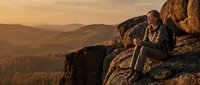 A young woman with medium skin tone sits on a rocky mountain outcrop at sunset, gazing at expansive golden hills and valleys below. Outdoor adventure and travel concept.