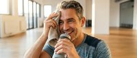 Mature man with gray hair and stubble, wearing casual dark t-shirt, holding microphone while speaking in bright studio or gym space. Suitable for motivational content, coaching, training, or presentation materials.