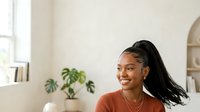 Young woman with dark skin tone and long black braided hair wearing a rust-colored sweater and gold necklace, smiling indoors with plants and shelving visible in background.