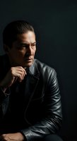 Middle-aged man with East Asian ethnicity wearing black leather jacket, contemplative pose with hand near face against dark background, suitable for corporate portraits and professional headshots.