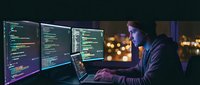 Young male programmer coding on multiple monitors in dark office environment with city lights background at night.