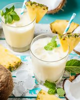 Fresh pineapple coconut smoothie with mint garnish and ice cubes on turquoise background, tropical summer drink served in glasses with fruit slices.