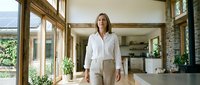 Middle-aged woman with shoulder-length blonde hair, Caucasian ethnicity, wearing white blouse and beige trousers, standing in modern home interior with large windows and natural light, suitable for lifestyle and real estate marketing.