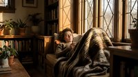 Young woman with curly hair relaxing on cozy window seat with plaid blanket in sunlit library room with bookshelves and plants for home comfort lifestyle.