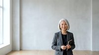Mature woman with white hair, Caucasian ethnicity, wearing dark blazer and black top, standing confidently in modern office space with minimalist decor and natural light from window.