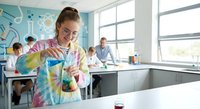 Young female student in tie-dye shirt conducting chemistry experiment with colorful liquid in laboratory classroom setting with classmates and teacher.
