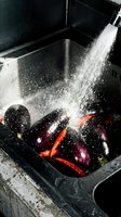 Fresh eggplants and red chili peppers being rinsed under running water in a black kitchen sink, with water droplets splashing. Food preparation and washing vegetables.