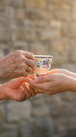 Elderly hands with wedding ring passing delicate floral teacup to younger hands in warm sunlight against stone wall background for family bonding concepts.