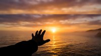 Hand reaching toward golden sunset over ocean waves with dramatic cloudy sky creating peaceful moment of connection with nature and hope.