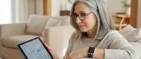 Mature woman with gray hair and glasses using tablet computer while relaxing on sofa at home, wearing smartwatch and cozy sweater in modern living room.