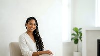 Young woman with dark skin tone and long braided hair wearing a white blazer, seated indoors with a potted plant visible in background. Suitable for corporate, business, and professional lifestyle content.
