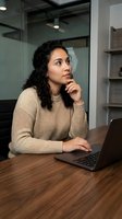 Young woman with long dark curly hair and medium skin tone working at wooden desk with laptop, wearing beige sweater, hand on chin in thoughtful pose, modern office setting with glass partitions.