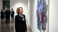Middle-aged Caucasian woman in black blazer admiring abstract artwork at contemporary art gallery exhibition with visitors in background.