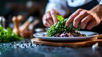 Professional chef garnishing gourmet dish with fresh microgreens and herbs on ceramic plate, close-up view of hands plating fine dining meal in restaurant kitchen.