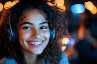 Young mixed race woman with curly hair wearing headphones smiles warmly at camera against blurred bokeh lights background, expressing joy and relaxation.