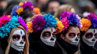 Women wearing traditional Day of Dead face paint and colorful flower crowns in vibrant blue, purple, and orange stand together during cultural celebration.