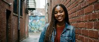 Young African American woman with braided hair smiling confidently in urban brick alley wearing denim jacket for lifestyle and portrait photography.