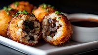 Deep fried rice balls filled with mushrooms and melted cheese, garnished with fresh herbs, served on white plate with dipping sauce, close up view of arancini.