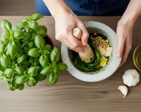 Woman preparing fresh pesto sauce in stone mortar with basil leaves and garlic on wooden kitchen counter for homemade Italian cooking and culinary preparation.