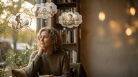 Middle-aged Caucasian woman with curly hair sitting thoughtfully in cozy cafe with thought bubbles showing memories of family moments and relationships.
