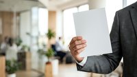 Businessman in dark suit holding blank white card in modern office environment with blurred colleagues and green plants in background, suitable for corporate communication and business messaging.