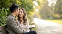 Happy young couple sitting on park bench sharing intimate moment during golden hour sunset with soft natural lighting and blurred background foliage.