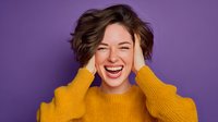 Young Caucasian woman laughing with hands on head wearing yellow sweater on purple background expressing joy and happiness for lifestyle content.