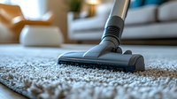 Close-up view of vacuum cleaner head cleaning light gray carpet with visible dust particles in modern living room interior with sofa and armchair in background.
