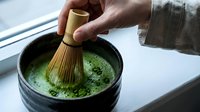 Caucasian male hand whisking matcha powder in ceramic bowl with bamboo whisk, preparing traditional green tea ceremony beverage for culinary and wellness content.
