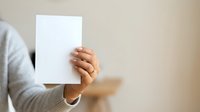 Woman holding blank white paper card in hand against neutral beige background, suitable for mockups, advertising, and promotional designs.