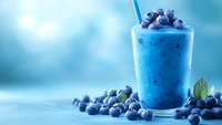 Fresh blueberry smoothie in glass with straw topped with berries on blue background for healthy breakfast drink concept.