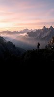 Silhouetted hiker stands on rocky mountain peak at sunrise, gazing over misty alpine valley with dramatic jagged peaks and golden sky. Adventure and exploration concept.