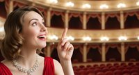 Woman in red evening gown with pearl necklace gestures thoughtfully inside ornate theater auditorium with golden balconies and warm lighting, suitable for entertainment and cultural event concepts.