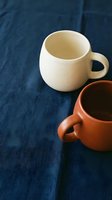 Two handmade ceramic coffee mugs in cream and terracotta colors on dark blue linen fabric background for kitchen decor and artisan pottery display.