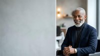 Senior African American male professional in dark blazer and turtleneck, smiling confidently in modern office setting with warm lighting, suitable for corporate, business, and leadership concepts.