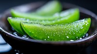 Fresh green aloe vera plant slices with water drops on dark ceramic plate, macro photography showing natural gel texture and moisturizing properties.
