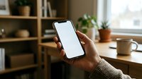 Person holding smartphone with blank white screen in modern home office workspace with wooden desk, shelving, plants and natural window light.