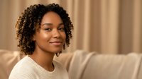 Young African American woman with curly hair smiling warmly while relaxing on beige sofa in cozy living room interior for lifestyle and wellness content.