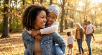 Happy African American mother carrying smiling toddler daughter on back in autumn park with golden sunlight and families enjoying outdoor activities.