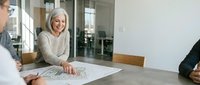 Senior businesswoman with gray hair smiling during office meeting, reviewing documents and plans with colleagues in modern corporate conference room.