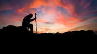 Silhouette of male hiker with trekking pole resting on rocky mountain summit against dramatic orange and pink sunset sky for adventure and outdoor concepts.