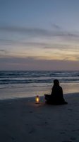 Silhouette of person meditating on beach at sunset with glowing lantern, peaceful ocean waves and colorful twilight sky creating serene atmosphere.